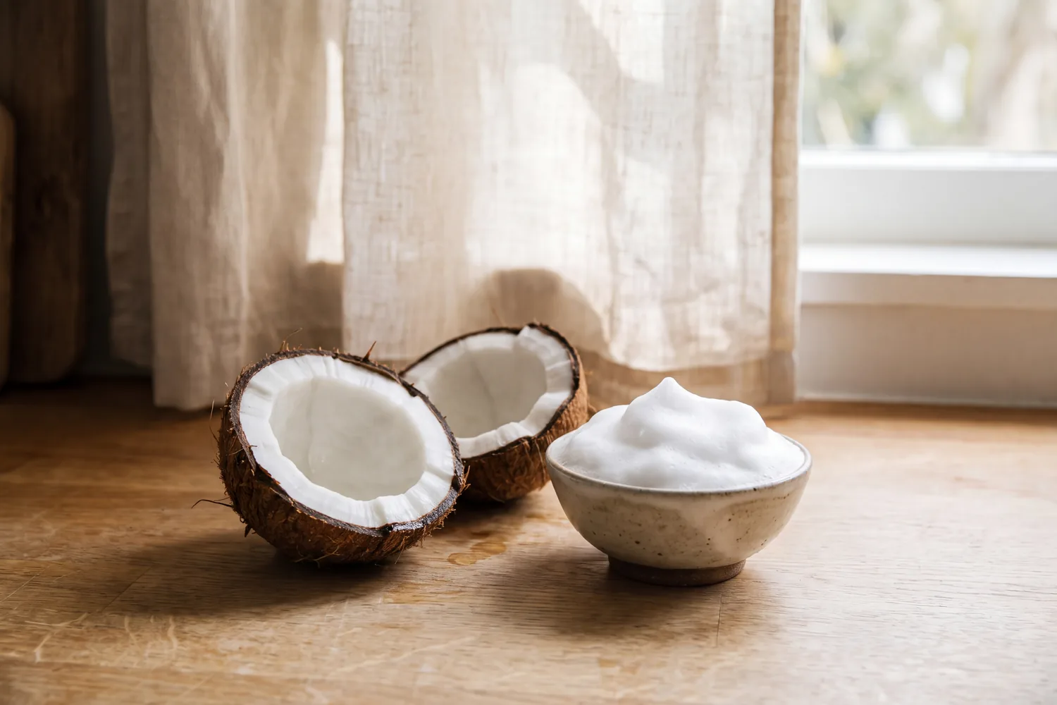 A coconut cracked in half beside a small ceramic bowl of white foam on an oak counter.